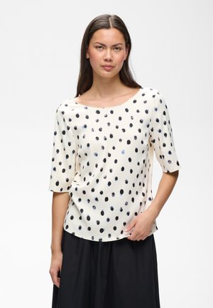 Femme aux longs cheveux foncés portant un haut blanc à pois noirs et des manches arrivant au coude, assortie d'une jupe noire, debout sur fond blanc.