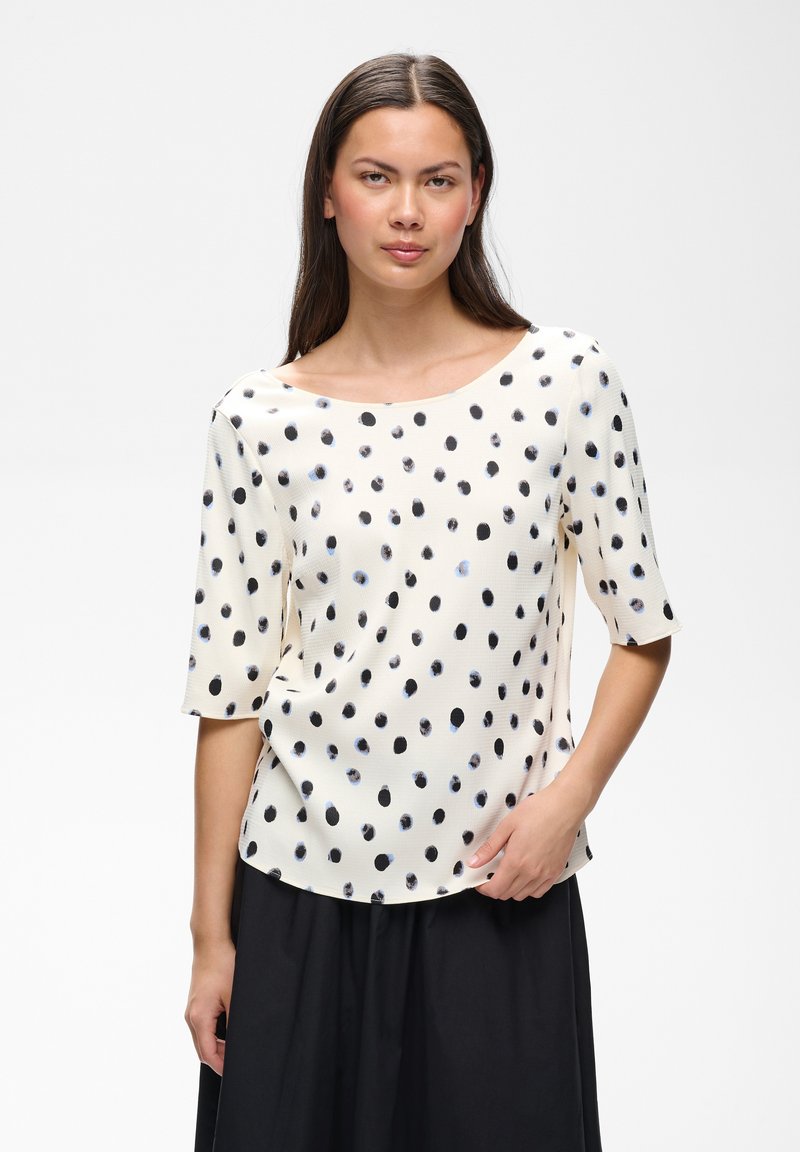 Femme aux longs cheveux foncés portant un haut blanc à pois noirs et des manches arrivant au coude, assortie d'une jupe noire, debout sur fond blanc.