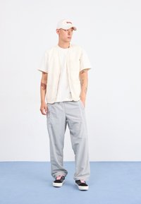 Gilet beige chiaro sopra una t-shirt bianca, abbinato a pantaloni grigi e sneakers nere con lacci rosa. Design semplice, look casual, dettagli minimi.