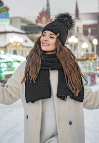 Black knit beanie with a fur pom-pom, paired with a textured black scarf. The model wears a cream-colored fleece coat in a snowy setting.