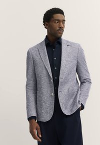 Hellgrauer strukturierter Blazer mit zwei vorderen Taschen und einem Knopfverschluss, getragen über einem dunkelblauen Hemd. Dezente Akzente unterstreichen das Design.