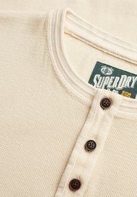 Cremefarbenes, strukturiertes Henley-Shirt mit drei dunklen Knöpfen und einem grünen Superdry-Markenlabel an der Innenseite des Kragens.