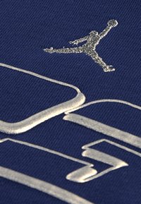 Granatowa koszulka z materiału z wyszytym srebrnym logo Jumpman i detalem tekstowym. Gładka struktura z lekko błyszczącym wykończeniem.