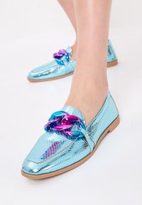 Pantofole metalice albastre cu textură de reptilă, prezentând o curea multicoloră ca accent. Talpă plată și design cu vârf ascuțit.