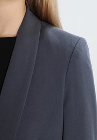 Grå blazer med en slät textur, skräddarsydd passform och vassa revers, delvis visad över en svart topp. Inga synliga knappar eller metalldelar.