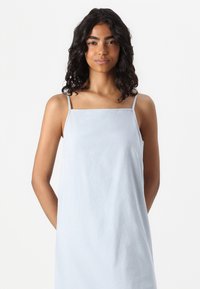 Ανοιχτό μπλε φόρεμα χωρίς μανίκια με λεπτές τιράντες, τετράγωνη λαιμόκοψη και χαλαρή εφαρμογή. Μαλακό, υφασμάτινο ύφασμα χωρίς σχέδια ή λεπτομέρειες.