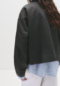 Veste noire surdimensionnée à texture lisse, dotée d'un col côtelé et de fermetures à pression, portée sur une chemise rayée bleu clair.