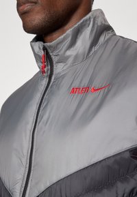 Detailný záber osoby v šedej a čiernej bunde s červeným logom "ATLETI" a Nike Swoosh na hrudi, zapnutou až po golier.