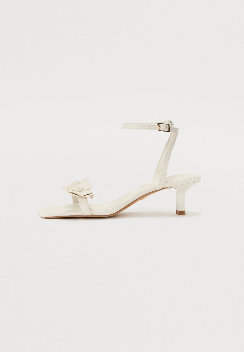 Bele sandale z belimi petami, kvadratnim nosom in majhno blok peto. Imajo nastavljiv trakom okrog gležnja ter dekorativni cvetlični poudarek na sprednjem delu.
