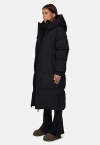 Długi czarny puchowy płaszcz z wysokim kołnierzem i kapturem, posiadający zamek błyskawiczny, boczne kieszenie oraz pikowaną konstrukcję. Miękki, izolowany materiał.