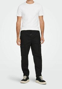 Černé cargo kalhoty s několika bočními kapsami, elastickým pasem a volným střihem, doplněné o jednoduché bílé tričko s krátkým rukávem a černé tenisky.