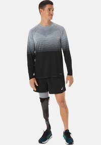 Langärmliges graues und schwarzes Sportshirt mit Farbverlaufsmuster, kombiniert mit schwarzen Shorts. Mit einer Prothesenbeine und blauen Sportschuhen.