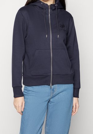 Person iført marineblå hættetrøje med lynlås, snore og forlommer, kombineret med lyseblå jeans, stående foran en ensfarvet baggrund.