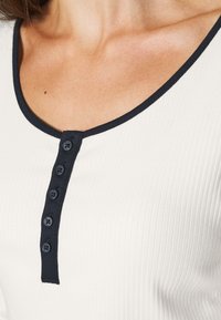 Pruhovaný bílý top s námořnickým kulatým výstřihem a knoflíkovou légou se šesti tmavými knoflíky. Tkanina má mírnou strukturu.