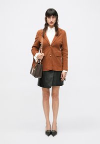 Barna velúrblézer, fehér blúz, fekete bőr szoknya és hegyes orrú fekete magassarkú. Barna táska mintás díszítéssel. Bojtokkal díszített táskafüggő.