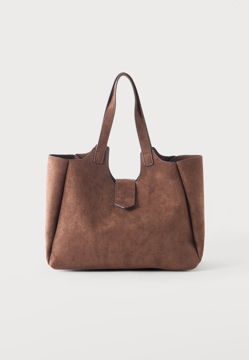 Sac cabas en daim marron avec deux poignées et une petite fermeture à rabat avant sur un fond clair uni.