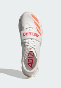 Basket de sport blanche avec des accents orange, tige en tissu texturé et strap Velcro. Comprend des lacets et un logo rouge proéminent sur la languette.