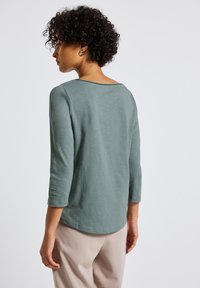 Helles grünes Langarmoberteil mit weitem Bootsausschnitt, weicher Textur und abgerundetem Saum. Das Modell trägt beige Hosen und zeigt die Rückansicht.