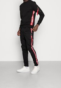 Man som bär en svart sweatshirt och byxor med röda "REMOVE BEFORE FLIGHT"-ränder och vita sneakers, står mot en vit vägg.