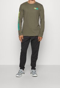 Man in een olijfgroene langslavende shirt met de tekst "Project Rock" en zwarte jogger pants met grijze en witte sneakers.
