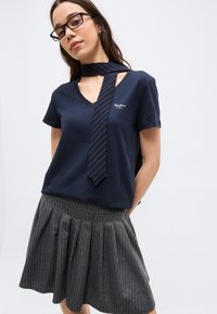 Γυναίκα που φοράει μαύρα γυαλιά, μπλε σκούρο T-shirt με V λαιμόκοψη και κείμενο, ένα μακρύ στενό ριγέ μαντίλι και μια γκρι ριγέ πιετέ φούστα.