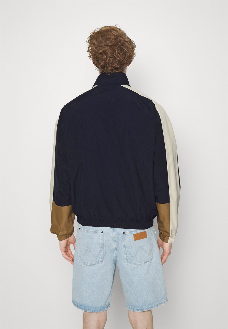 Marineblauwe jas met crèmekleurige en bruine accenten, elastische manchetten en zoom, gestructureerd materiaal, achterkant, gedragen over lichte denimshorts.