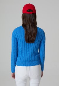 Blauer Kabelstrickpullover mit langen Ärmeln und geripptem Saum. Kombiniert mit weißen Jeans und einer roten Baseballmütze, von hinten betrachtet.