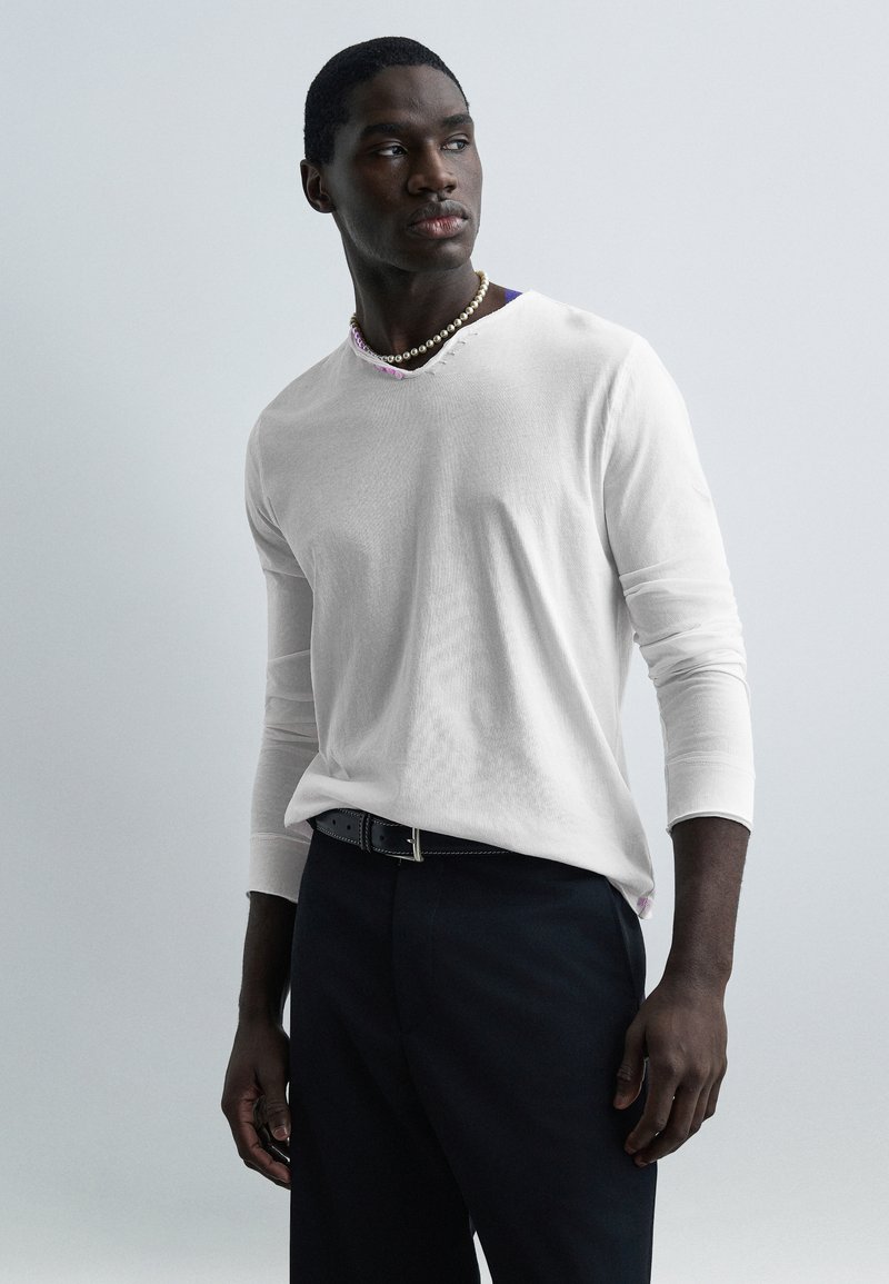 Άνδρας με άσπρο πουκάμισο μακρυμάνικο, μαύρο παντελόνι, μαύρη ζώνη και μαργαριταρένιο κολιέ, στέκεται μπροστά σε ανοιχτό γκρι φόντο, κοιτάζοντας προς το πλάι.