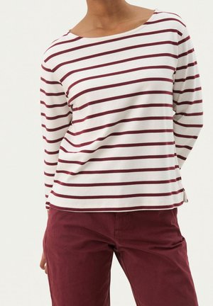 Personne portant une chemise blanche à manches longues avec des rayures horizontales bordeaux et un pantalon bordeaux uni, debout les mains dans le dos.