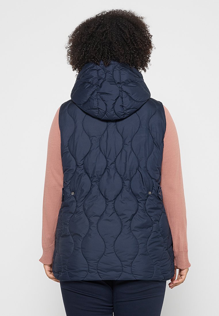 Veste matelassée marine avec capuche, présentant une texture ondulée et des boutons-pression sur les côtés, portée par-dessus un haut à manches longues rose.