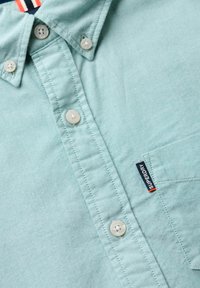 Camicia button-up verde chiaro realizzata in tessuto testurizzato con colletto, presenta bottoni bianchi e un piccolo tag con il marchio sul petto.