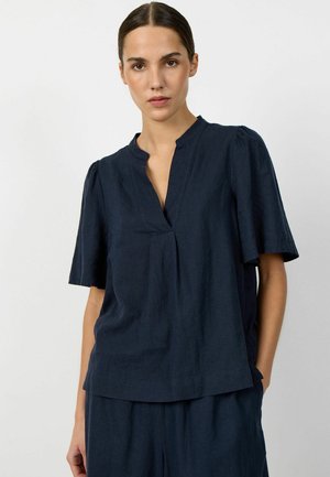 Frau mit zurückgebundenem dunklem Haar trägt eine marineblaue Kurzarmbluse mit V-förmigem Ausschnitt und passende marineblaue Hose.