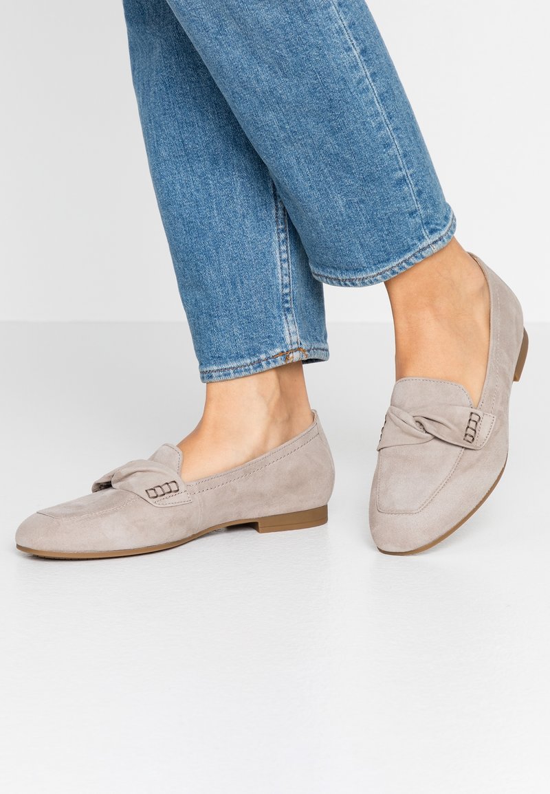Pieds portant des mocassins en daim gris clair avec un détail de nœud, associés à un jean en denim bleu, debout sur un sol clair.