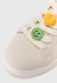 Beige sneaker with a smooth texture, white laces, and three colorful decorative lace charms: a green smiley face, a yellow heart, and an orange accent.