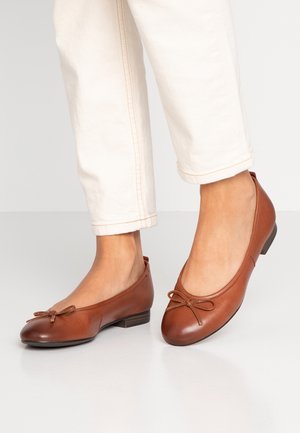 Ballerines en cuir marron avec un bout rond et un détail de nœud décoratif. Talon bas et texture lisse, associées à un pantalon de couleur claire.
