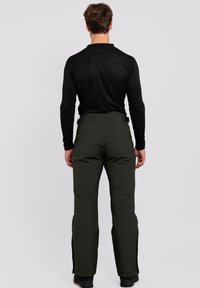 Pantalones de nieve verdes con cremalleras laterales, una cintura ajustada y un tejido duradero. Se llevan con una camiseta de manga larga negra, mostrando una textura suave.