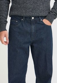 Jeans de mezclilla azul oscuro con corte recto, que presentan un diseño clásico de cinco bolsillos, herrajes en plata y una ligera textura.
