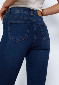 Tmavě modré džíny z denimu s přiléhavým střihem, dvěma zadními kapsami a koženou nášivkou na pase. Hladká textura, minimální detaily švů.