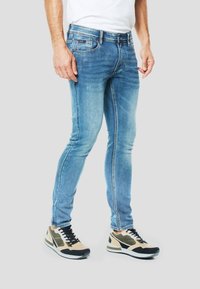 Jeans slim fit azzurro chiaro con lievi segni di usura sulle cosce, abbinati a scarpe da ginnastica beige e nere con design a rete e lacci.