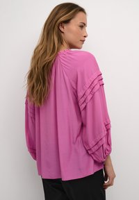 Blusa rosa, ampia e morbida, con maniche a sbuffo, scollatura arricciata e una texture soffice. Il retro presenta dettagli a strati.