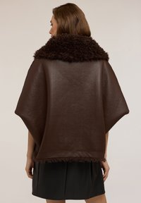 Žena v tmavě hnědém koženém plášti s tlustým texturovaným límcem a lemem, v kombinaci s černou sukní, zobrazená zezadu.