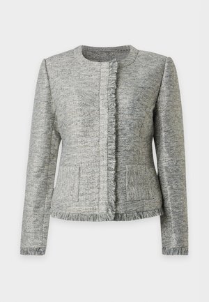 Mujer con cabello rubio liso que lleva una chaqueta gris texturizada, pantalones cortos de cuero negros hasta la rodilla y zapatos slingback negros de tacón.