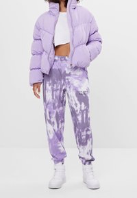 Liliowy puchowy kurtka z pikowanymi sekcjami; krótkie białe top; spodnie dresowe w fioletowy tie-dye z teksturowanym wzorem; białe sneakersy typu high-top.