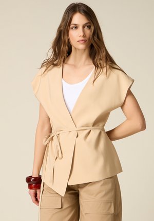 Donna che indossa un top beige senza maniche avvolgente con cintura da annodare, canotta bianca, pantaloni cargo beige e due braccialetti rossi spessi al polso sinistro.