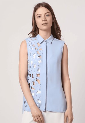 Blusa sin mangas de color azul claro con cierre de botones en la parte frontal, que presenta un diseño de corte floral en el lado izquierdo y una textura de tela suave.