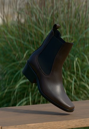 Bota de cuero marrón hasta el tobillo con un diseño elegante, paneles laterales elásticos, punta afilada y un tacón bajo, en un fondo verde.