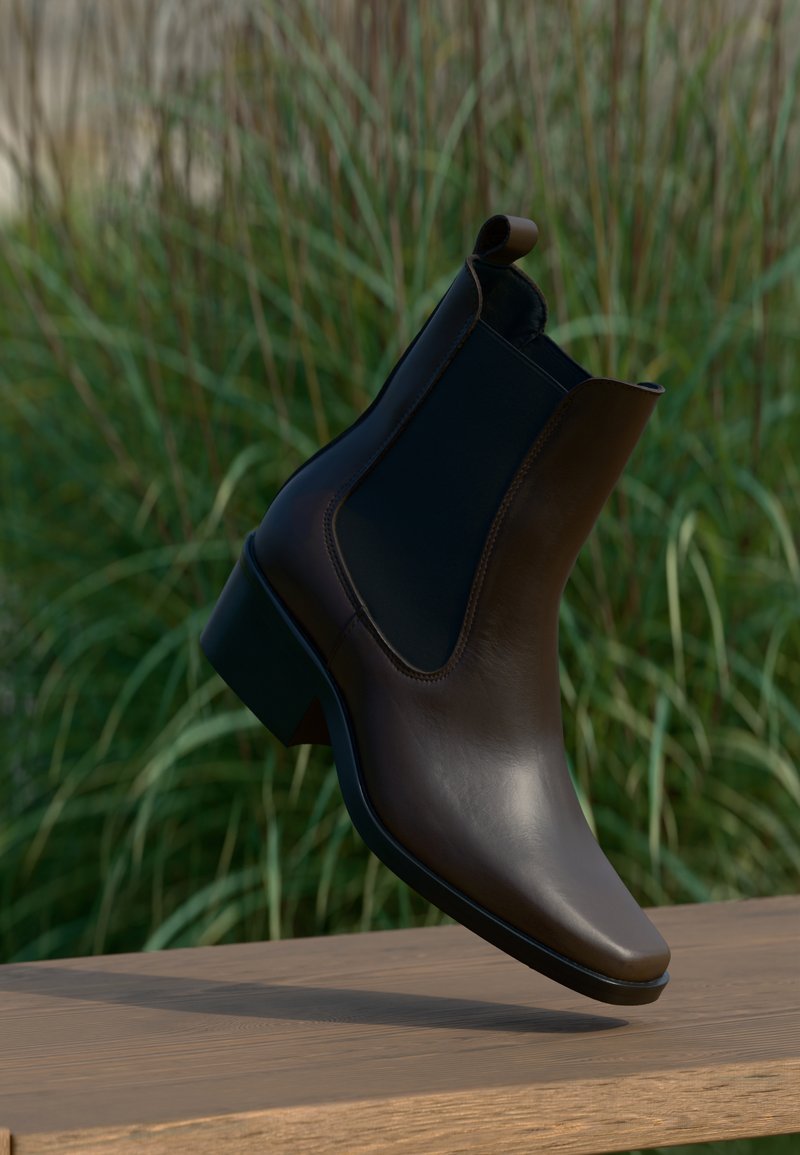 Καφέ δερμάτινη μποτάκι έως τον αστράγαλο με κομψό σχέδιο, ελαστικές πλευρές, μυτερή μύτη και χαμηλό τακούνι, σε φόντο πράσινο.