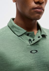 Κοντινό πλάνο ενός ατόμου που φοράει ένα πράσινο πουκάμισο με γιακά, μαύρα κουμπιά και μαύρο οβάλ λογότυπο στο στήθος.