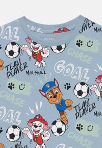 Gaiši zila īsām piedurknēm T-krekls ar karikatūru suņiem, kas spēlē futbolu, ar tekstiem «MĒRKWN» un «IZCILIE KOMPONENTS», kā arī pēdu nospiedumu motīviem.