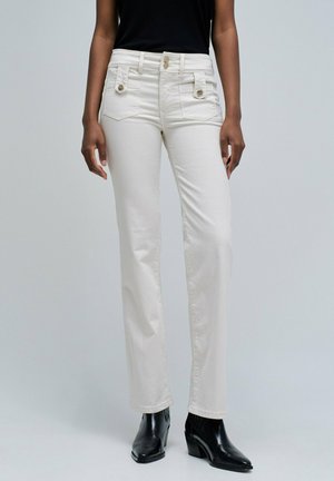 Personne portant un pantalon blanc taille haute et droit avec des poches avant boutonnées, assorti à des bottes noires à talons aiguilles et un haut noir.